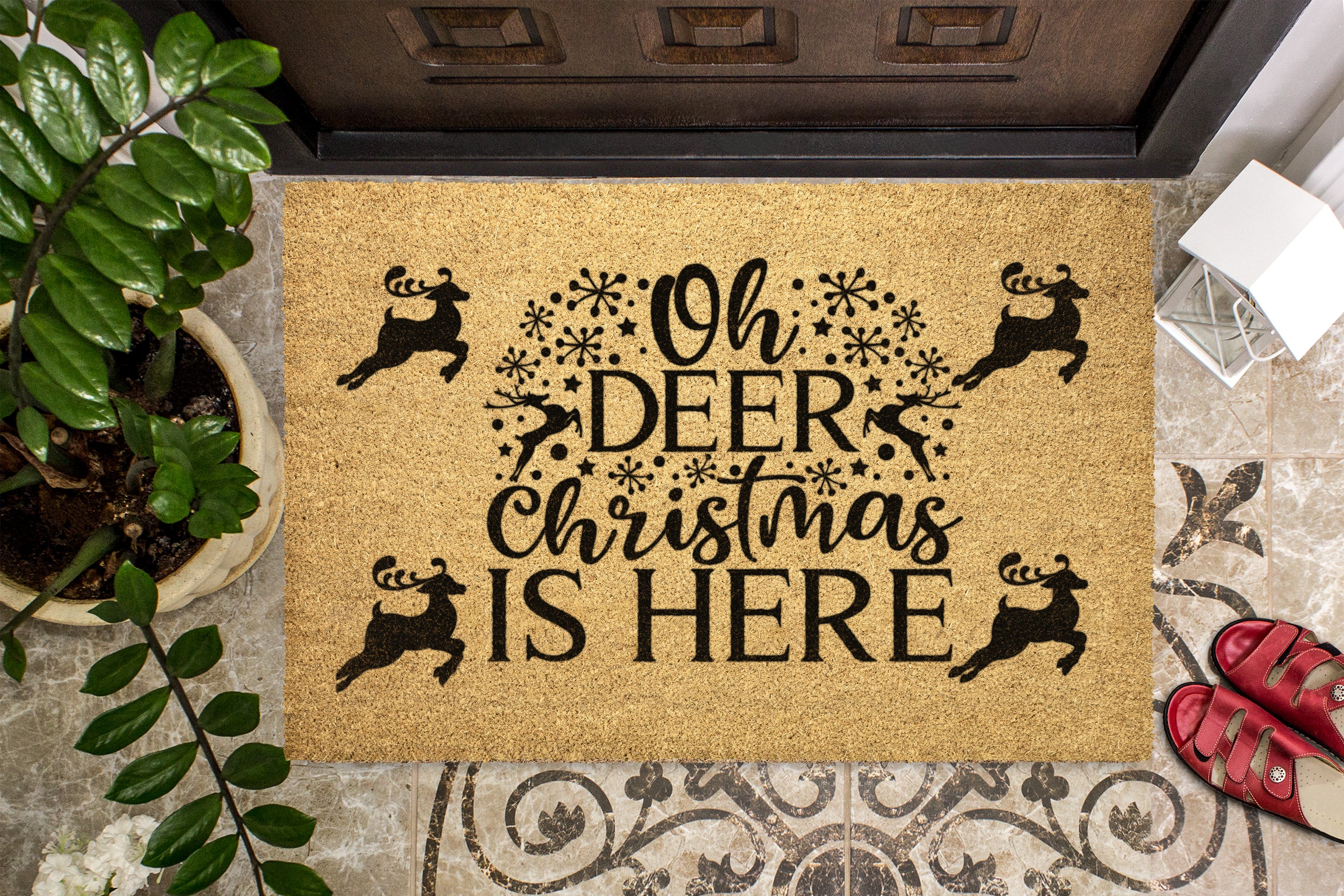 A rectangular coir doormat with the phrase 'Oh DEER Christmas IS HERE' in black lettering, surrounded by black reindeer silhouettes and snowflake motifs. The doormat is staged on a tiled entryway floor next to a potted plant, a door threshold, and a pair of red sandals.
