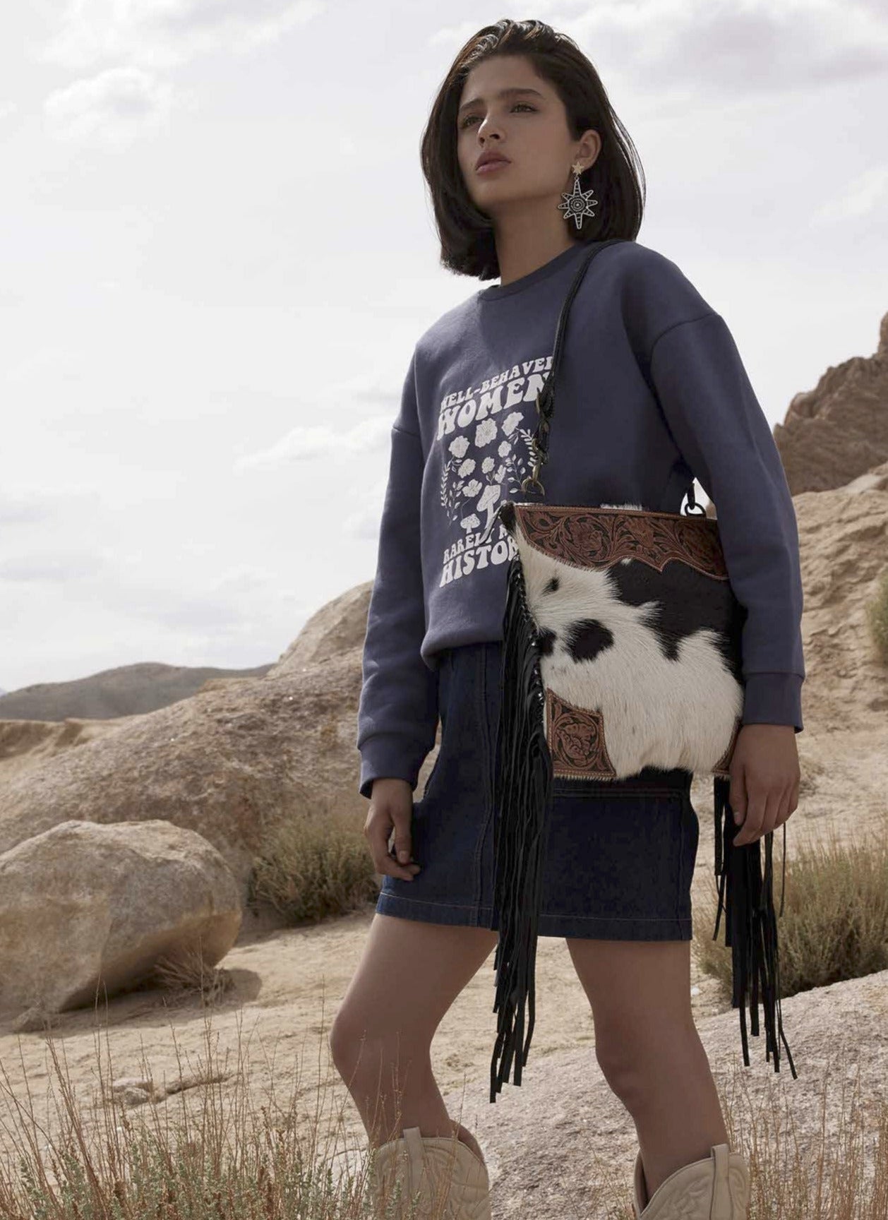 Woman standing in a desert landscape holding a cow-patterned bag
